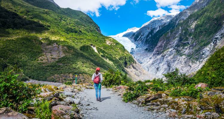 Una excursionista sola camina por un sendero en el valle hacia el glaciar distante, acurrucado entre colinas verdes exuberantes y el cielo azul.