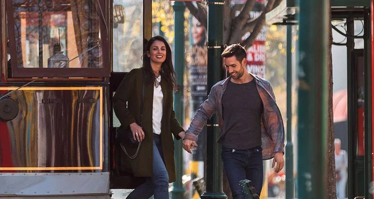 Pareja sonriente bajando de un tranvía urbano vintage a una calle soleada bordeada de columnas verdes.