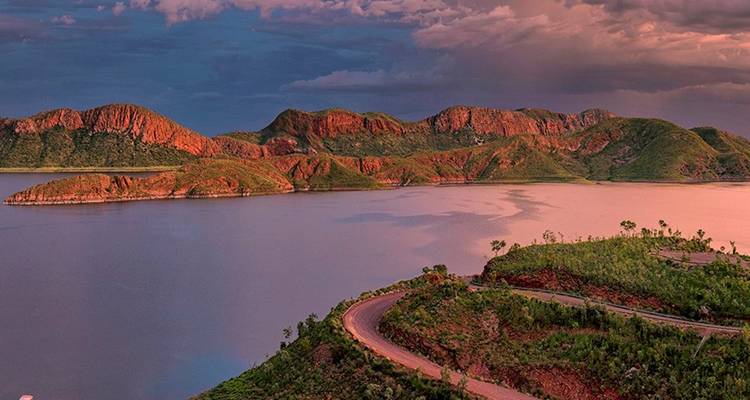 Lago sereno bordeado por acantilados rojos y colinas verdes bajo un cielo de atardecer en tonos pastel.