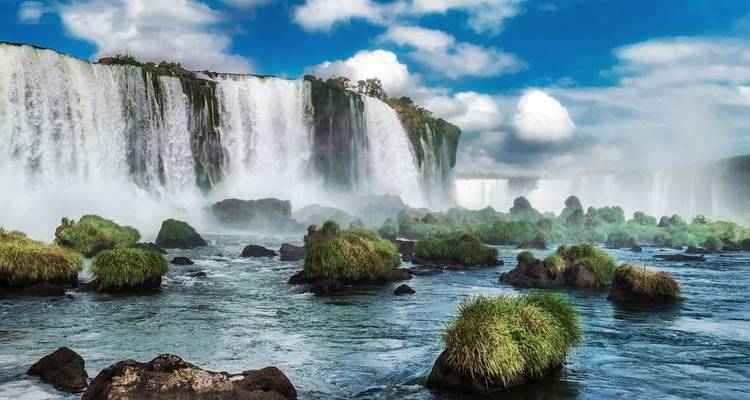 Puissant rideau des chutes d'Iguazu se déversant dans une rivière brumeuse entourée d'une végétation luxuriante sous un ciel bleu éclatant avec des nuages épars.