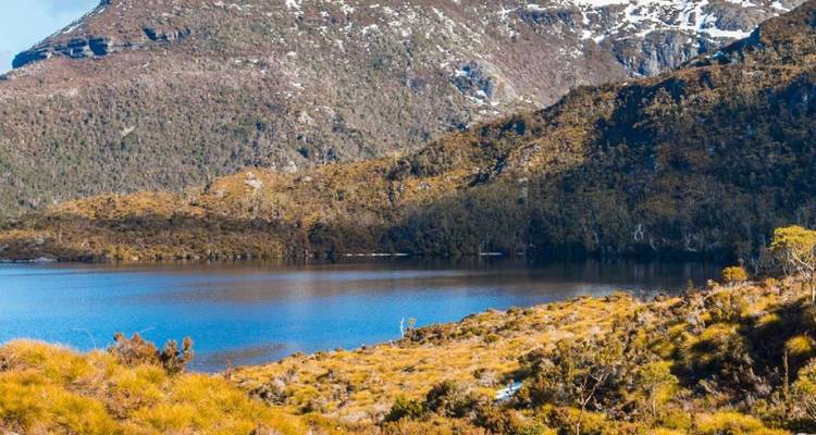 Lago alpino azul cristalino que refleja picos escarpados salpicados de nieve y laderas boscosas en Tasmania.