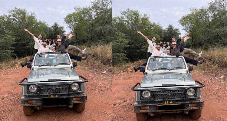 Eine fröhliche Gruppe posiert auf einem Safari-Jeep auf einer roten Lehmstraße, umgeben von trockener Waldvegetation.