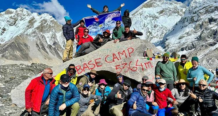 Group of people posing with a rock marked 'Base Camp 5364m' and snowy mountains.