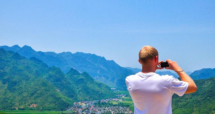 Male traveller photographs a broad valley of green mountains and a village far below on a sunny day.