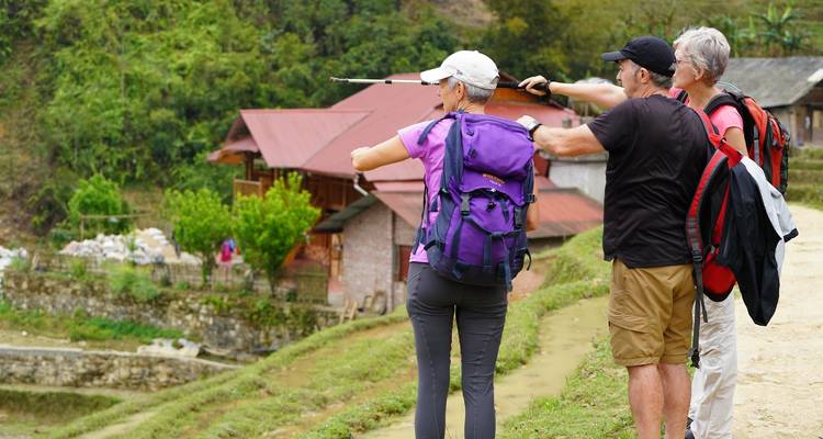 Three trekkers consult a trekking pole and point toward village houses in a valley.