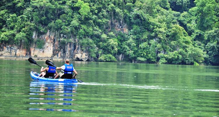 Two travelers paddling a blue kayak on a calm green lake framed by limestone cliffs and thick jungle.