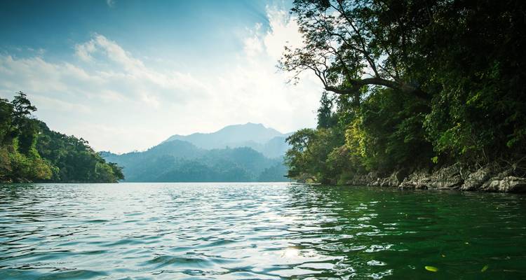 Serene emerald lake surrounded by dense tropical forest and distant misty mountains.