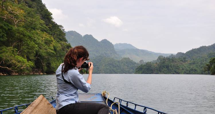 Young woman seated at the bow of a small boat taking photos of misty, forested mountains across the lake.