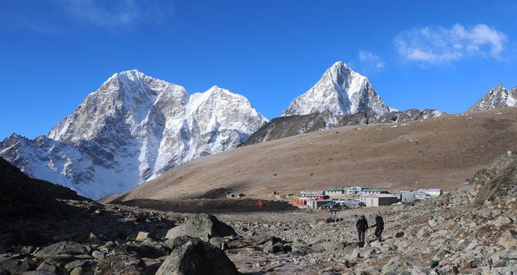 Dos trekistas caminan hacia un asentamiento de gran altura respaldado por dramáticos picos del Himalaya cubiertos de nieve.