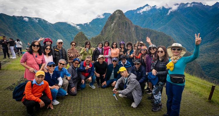 Un gran grupo de turismo posa alegremente en la terraza con vistas a las ruinas de Machu Picchu y los picos de los Andes envueltos en nubes.