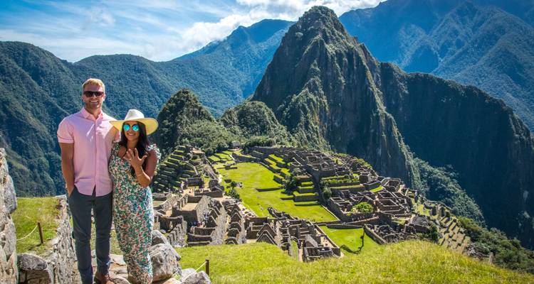 Una pareja sonriente se encuentra de pie en una terraza de piedra inca con una vista panorámica de la ciudadela de Machu Picchu abajo.