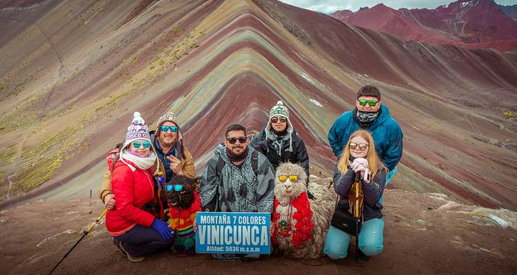 Los viajeros abrigados con ropa de invierno posan ante las rayas de colores del Arco Iris de la Montaña Rainbow Mountain sosteniendo un cartel.