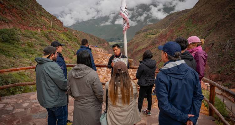 Un guía habla con un pequeño grupo de turistas en un mirador sobre las salinas en terrazas en un valle montañoso.