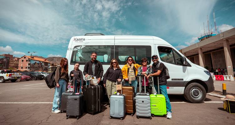 Viajeros con maletas coloridas posan junto a una furgoneta de tour blanca en un estacionamiento.
