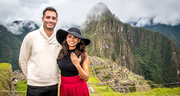 Una pareja comprometida sonríe con el dramático pico de Machu Picchu detrás de ellos, la mujer mostrando su anillo.