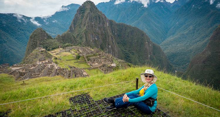 Una mujer se sienta en el pasto con las terrazas de Machu Picchu y el pico de Huayna Picchu asomándose detrás de ella.