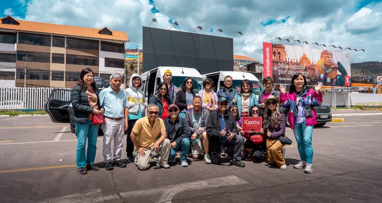 Un gran grupo de turismo posando frente a una furgoneta de pasajeros blanca en un estacionamiento de aeropuerto con montañas y vallas publicitarias de fondo.