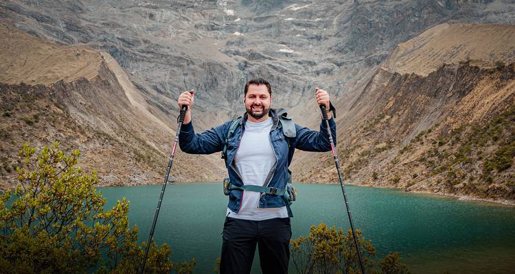 Un excursionista levantando triunfalmente bastones de trekking frente a un lago alpino de color turquesa rodeado de montañas rocosas escarpadas.