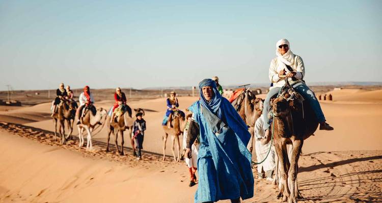 Kameelkaravaan geleid door lokale gids die gouden woestijnduinen oversteekt onder heldere blauwe hemel.