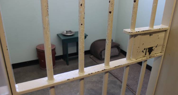Intérieur d'une cellule de prison avec des équipements de base, visible à travers des barreaux de fer.