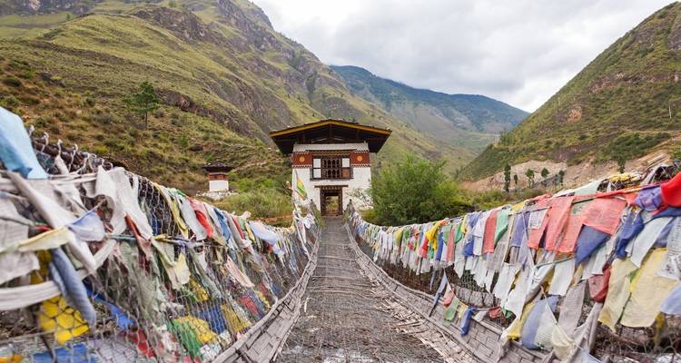 Een hangbrug versierd met kleurrijke gebedsvlaggen die naar een huis in de bergen leidt.