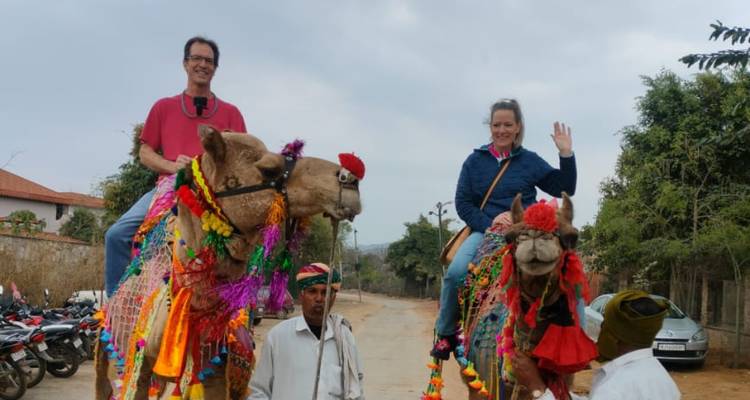 Touristen, die auf hell geschmückten Kamelen reiten, die von lokalen Führern entlang einer staubigen Dorfstraße geleitet werden.