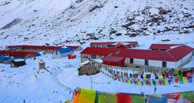 Grupo de alojamientos con techos rojos y banderas de oración ondeando en una cuenca himalaya nevada bajo picos escarpados.