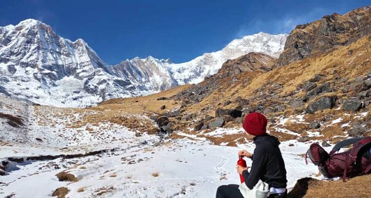 Trekker con gorro rojo descansando en un prado alpino nevado mirando hacia los imponentes picos blancos del Himalaya.