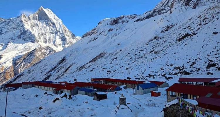 Refugios cubiertos de nieve dispersos a lo largo de una pendiente pronunciada del Himalaya bajo un dramático pico en forma de aleta de tiburón.