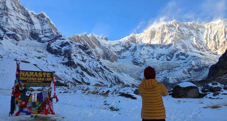 Un excursionista con chaqueta amarilla se para frente a un anfiteatro nevado gigante en el Campamento Base Annapurna junto a un colorido letrero de bienvenida.