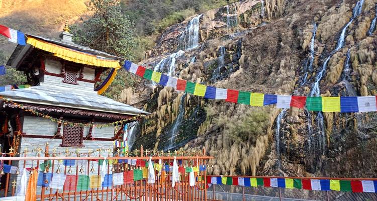 Pequeño santuario himalayo decorado con banderas de oración junto a cascadas que caen en un acantilado rocoso.