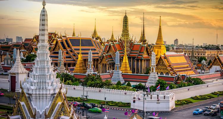 Las agujas doradas y los tejados coloridos del Gran Palacio de Bangkok brillan con los tonos cálidos del atardecer con el horizonte de la ciudad de fondo.