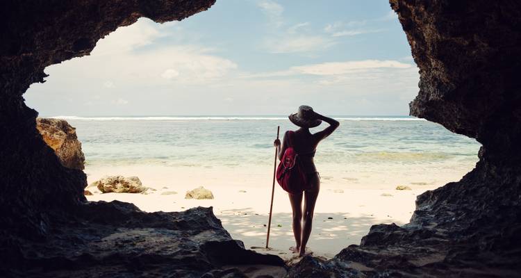 A lone traveller silhouetted in a rocky cave entrance looking out to a calm sandy beach and sea.