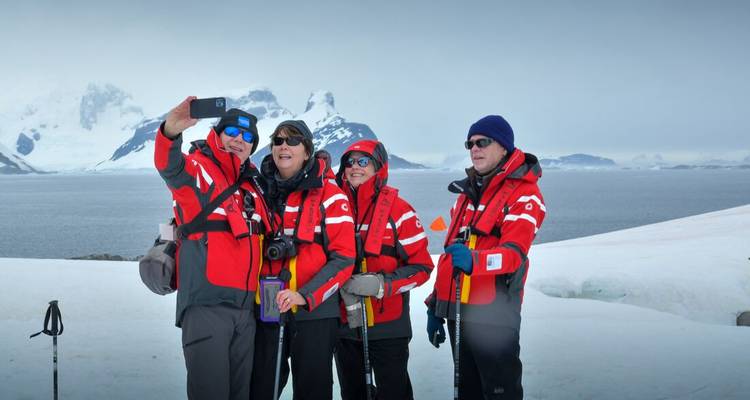 Vier expeditieleden in rode polaire jassen poseren voor een selfie op een besneeuwde Antarctische kustlijn met bergen op de achtergrond.