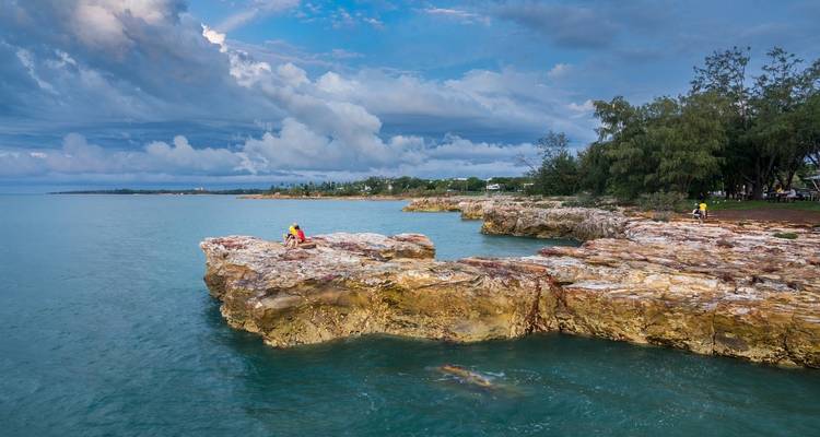 Turquoise sea lapping against layered coastal rock ledges with people sitting on the cliffs near tropical Darwin shoreline.