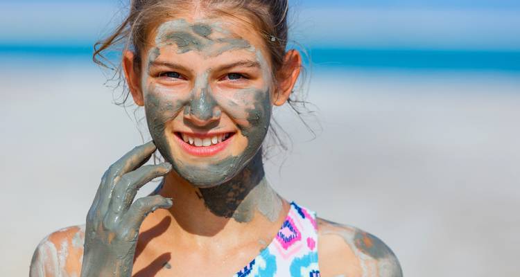 Persoon met modder op hun gezicht die glimlacht naar de camera bij een strand.