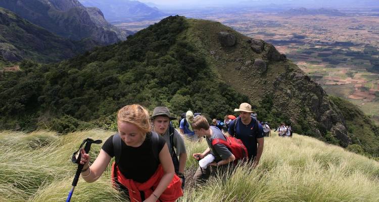 Groupe de randonneurs gravissant un sentier de montagne herbeux avec une vue panoramique en arrière-plan.