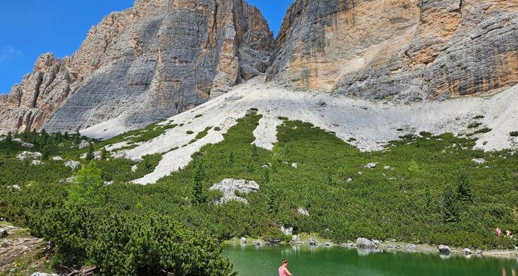 Un senderista solitario se para junto a un pequeño lago alpino verde bajo los imponentes acantilados grises de las Dolomitas.