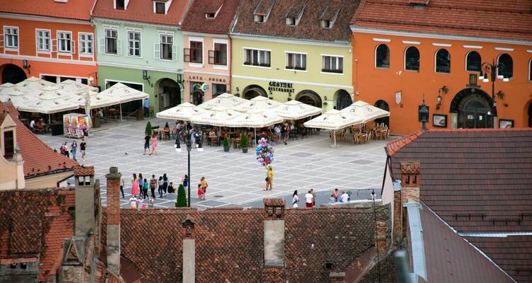 Vue aérienne d'une place médiévale colorée bordée de parasols de café et de bâtiments aux couleurs pastel.