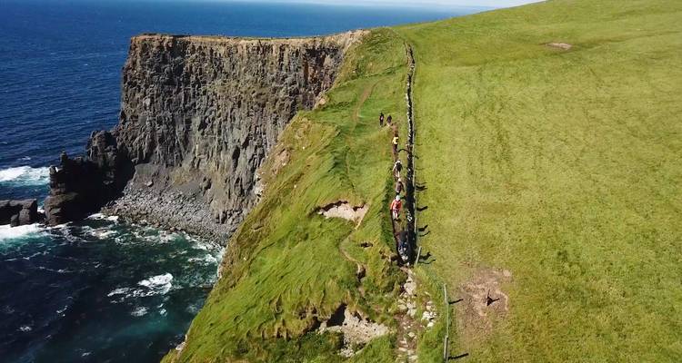 A group of people walking along the top of a grassy cliff above the sea.