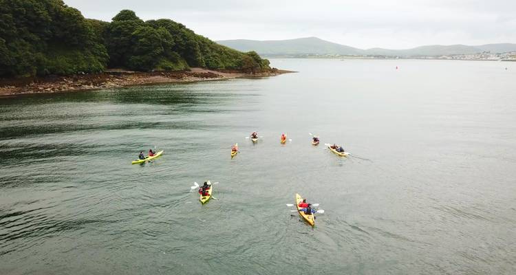 A group of people kayaking in a calm coastal area with hills in the background.