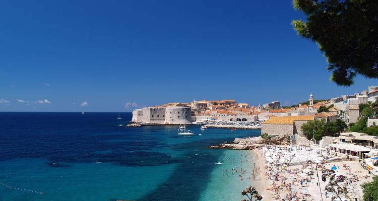 Vue panoramique de la vieille ville fortifiée de Dubrovnik s'avançant dans l'Adriatique azurée avec une plage animée au premier plan.
