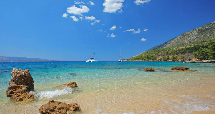 Baie aux eaux cristallines turquoise avec plage de sable, voiliers lointains et collines boisées sous un ciel éclatant.