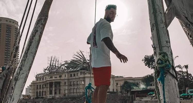 Man staand op een houten boot kijkend naar de camera met historische gebouwen op de achtergrond.