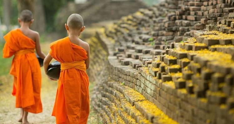 Zwei junge Mönche in orangefarbenen Roben gehen entlang einer alten Backsteinmauer.
