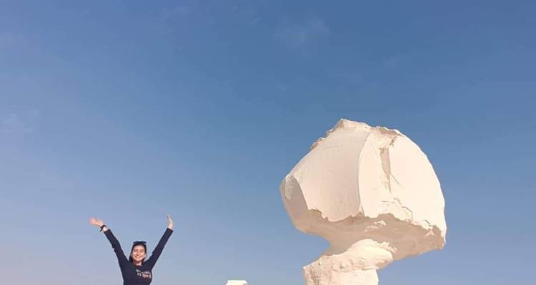 Een persoon met opgeheven handen naast een grote rots onder een heldere blauwe lucht.