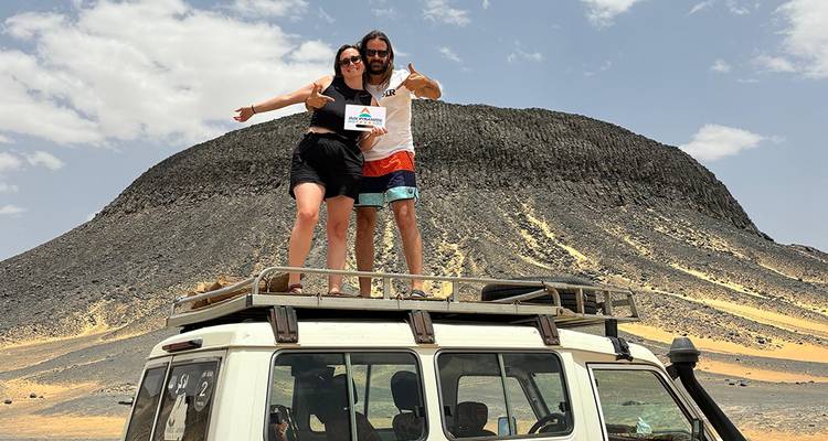 Two people standing on a vehicle roof in a desert landscape.