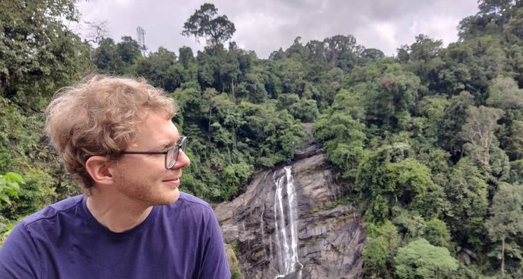 Der Reisende blickt über ein üppiges Dschungeltal auf einen hohen Wasserfall, der über dunklem Felsen hinabstürzt.