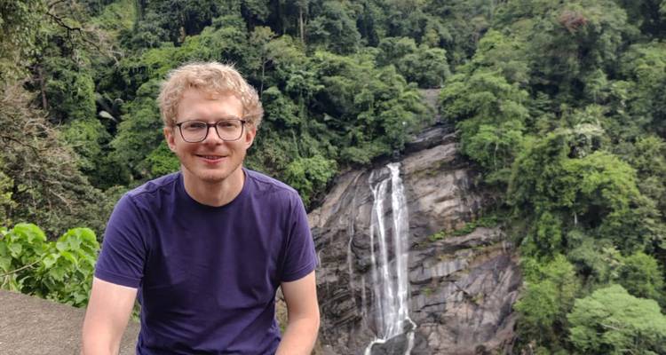Lächelnder Reisender sitzt vor einem üppigen Klippen-Wasserfall inmitten eines dichten Regenwaldes.