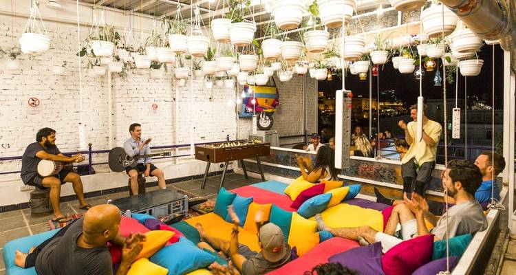 Hostel gemeenschappelijke ruimte met kleurrijke vloerkussens, gasten die muziek spelen en tafelvoetbal onder hangende planten 's nachts